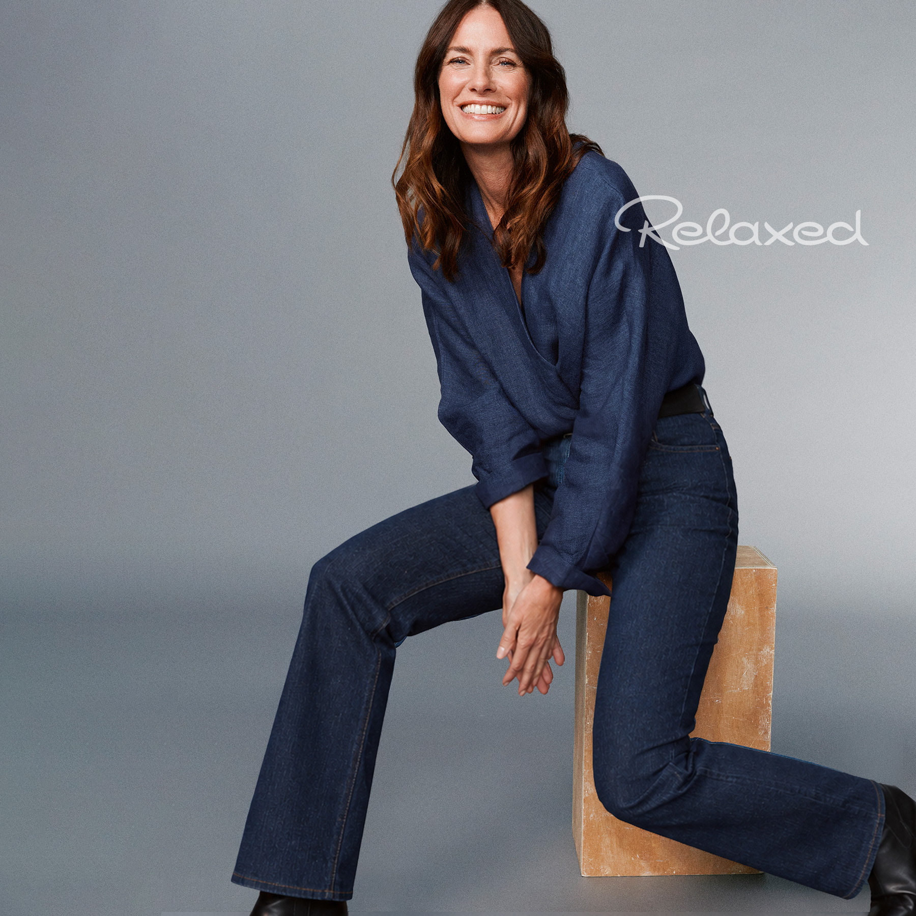 Smiling woman wearing a dark blue blouse and wide-leg jeans, seated on a wooden stool against a grey background – mobile hero teaser for the RELAXED brand and its Comfort Meets Style collection