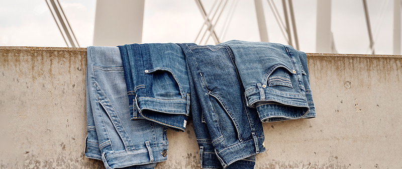 Jeans für jeden Tag. Und jeden Stil. Vier gefaltete TONI-Jeans in verschiedenen Waschungen auf Betonmauer drapiert.