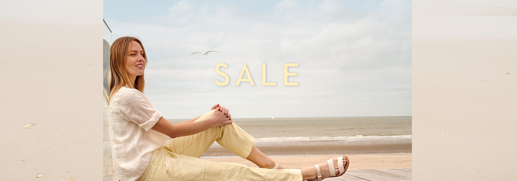 Woman in light-toned TONI outfit relaxing at the beach – summery sale teaser.