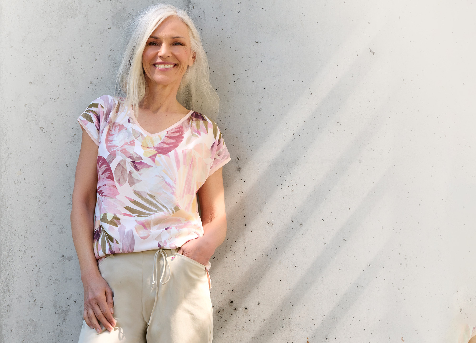 Smiling woman wearing a floral top and light-coloured trousers against a concrete wall – hero banner for the TONI Relaxed collection